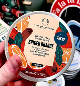 A short cylindrical silver metallic tin full of some creamy white body butter with a silver metallic lid and a white label that has The Body Shop Spiced Orange Body Butter written in dark green writing and some pictures of some large spherical Oranges on it, on a white background.