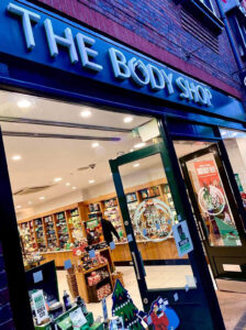 A large brown stone rectangular shaped building with a large rectangular dark green board over it's doors that has The Body Shop written in bold white writing on it, on a white background.