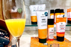 A tall cylindrical champagne flute full of some bright orange liquid in front of a collection of different shaped and coloured Glass Bottles, Plastic Tubs, and Plastic Tubes that all have The Body Shop Vitamin C written in bold dark green writing on them on some light wooden shelves.