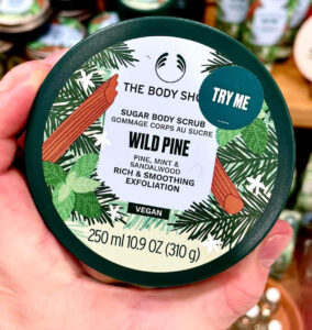 A short cylindrical silver metallic tin full of some creamy white body butter with a silver metallic lid and a white label that has The Body Shop Wild Pine Body Butter written in dark green writing and some pictures of some dark green pine trees on it, on a white background.