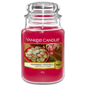 A tall cylindrical glass jar full of some bright red wax with a label that has Yankee Candle written in white writing, peppermint Pinwheels written in black writing, and a picture of some bright red and white swirled Peppermint Pinwheels on a circular black plate on it, on a white background.