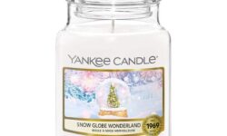 A tall cylindrical glass jar full of some creamy white wax with a label that has Yankee Candle written in white writing, Snow Globe Wonderland written in black writing, and a picture of a large dark green Christmas Tree and a silver circular snow globe on a white Table on it, on a white background.