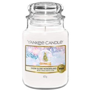 A tall cylindrical glass jar full of some creamy white wax with a label that has Yankee Candle written in white writing, Snow Globe Wonderland written in black writing, and a picture of a large dark green Christmas Tree and a silver circular snow globe on a white Table on it, on a white background.