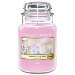 A tall cylindrical glass jar full of some pale pink wax with a label that has Yankee Candle written in white writing, snowflake kisses written in black writing, and some pictures of some white falling snowflakes on it, on a white background.