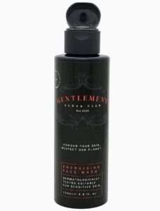 A tall cylindrical black plastic bottle with a black plastic lid and a black label that has Gentlemen’s Scrub Club Energising Face Wash written in bright red writing on it, on a white background.