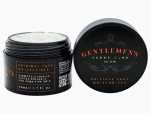 A short cylindrical black Plastic Tub full of some creamy white liquid that has some pieces of Cranberry Seeds running through it with a black Plastic Lid and a black label that has Gentlemen’s Scrub Club Exfoliating Face Scrub written in bright red writing on it, on a white background.
