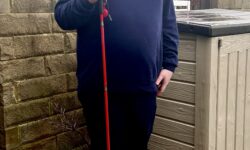 A tall bearded man wearing a navy blue crew neck jumper, a navy blue v-neck t-shirt, a pair of navy blue chino trousers and a pair of shiny red shoes holding a bright red cane stood against a dark wooden fence.