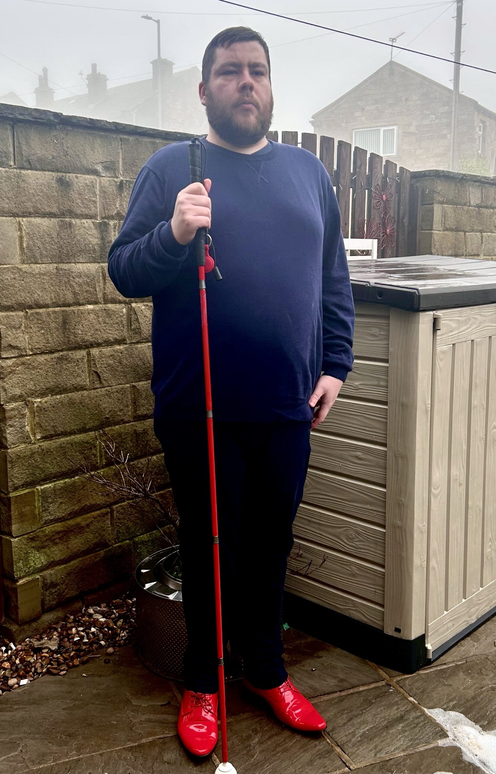 Outfit Of The Day | Jon Richardson The Knitwit Tour At The St. George’s Hall Bradford. A tall bearded man wearing a navy blue crew neck jumper, a navy blue v-neck t-shirt, a pair of navy blue chino trousers and a pair of shiny red shoes holding a bright red cane stood against a dark wooden fence.