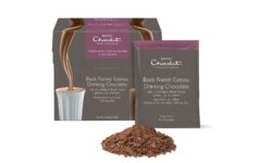 A tall rectangular dark grey cardboard box that has Hotel Chocolat Black Forest Gateaux Hot Chocolate written in bright white writing and a picture of some Hot Chocolate being poured into a short cylindrical cup on it next to a pile of some dark brown hot chocolate powder, on a white background.