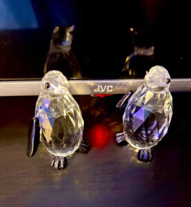 Two small black and white crystal penguins in front of a large rectangular black flatscreen Tv, on a black wooden cabinet.