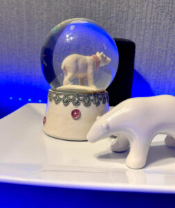 A large bright white spherical snow globe that has a polar bear stood on all four paws inside it next to a small white polar bear that’s stood on all four paws on top of a large rectangular white plate and is surrounded by some bright blue strip lights, on a black wooden cabinet.