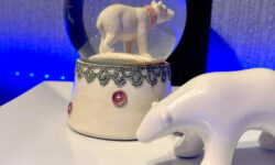 A large bright white spherical snow globe that has a polar bear stood on all four paws inside it next to a small white polar bear that’s stood on all four paws on top of a large rectangular white plate and is surrounded by some bright blue strip lights, on a black wooden cabinet.