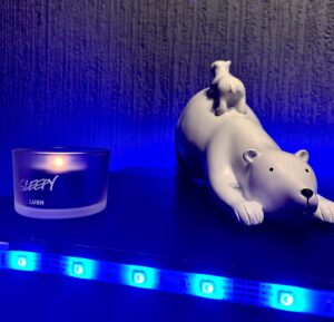 A large bright white polar bear that’s laid on it’s stomach and has a small bright white baby polar bear sat on it’s back behind a small cylindrical frosted glass Tea Light Holder that has a lit bright white Tea Light inside it, on a black wooden cabinet.