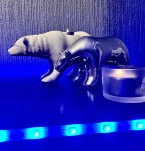 A large bright white Polar Bear Tea Light Holder that has a lit bright white Tea Light coming out of the top of it next to a medium silver Polar Bear behind a small cylindrical frosted glass Tea light holder that has a lit bright white tea light inside it, on a black wooden cabinet.