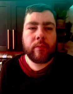 A tall bearded man wearing a black crew neck jumper stood against some dark navy kitchen cupboards.