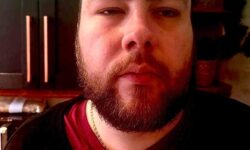 A tall bearded man wearing a black crew neck jumper stood against some dark navy kitchen cupboards.