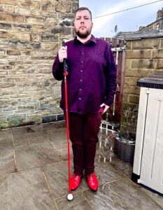 A tall bearded man wearing a dark red long sleeve oxford’ shirt, a dark red V-neck T-shirt, a pair of dark red chino trousers and a pair of shiny red shoes holding a long red cane stood against a dark wooden fence.