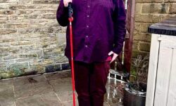 A tall bearded man wearing a dark red long sleeve oxford’ shirt, a dark red V-neck T-shirt, a pair of dark red chino trousers and a pair of shiny red shoes holding a long red cane stood against a dark wooden fence.
