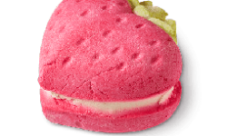 A bright Red Strawberry shaped Bubbleroon with a dark green stem and a cut In the middle of it that's filled with some bright white Cocoa Butter, on a white background.