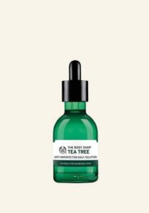 A short cylindrical brown glass bottle with a black rubber pipette lid and a bright white label that has The Body Shop Tea Tree Anti-Imperfection Daily Solution written in dark green writing on it, on a white background.
