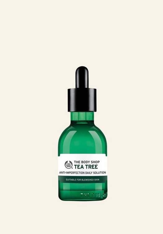 Using Another Three Leaves From The Same Tree | The Body Shop Tea Tree Range A short cylindrical brown glass bottle with a black rubber pipette lid and a bright white label that has The Body Shop Tea Tree Anti-Imperfection Daily Solution written in dark green writing on it, on a white background.
