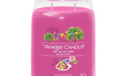 A tall cylindrical glass jar full of some bright pink wax with a label that has Yankee Candle written in white writing, Art In The Park written in black writing, and a picture of an art easel next to some bright green trees covered in some bright white bunting on it, on a white background..