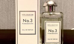 A tall rectangular clear glass bottle full of some clear liquid with a metallic silver lid and a white label that has Aldi Hotel Collection Pomegranate Noir Eau De Parfum written in bold black writing on it next to a tall rectangular white cardboard box that has Aldi Hotel Collection No.3 Pomegranate Noir Eau De Parfum written in bold black writing on it, on a white background.