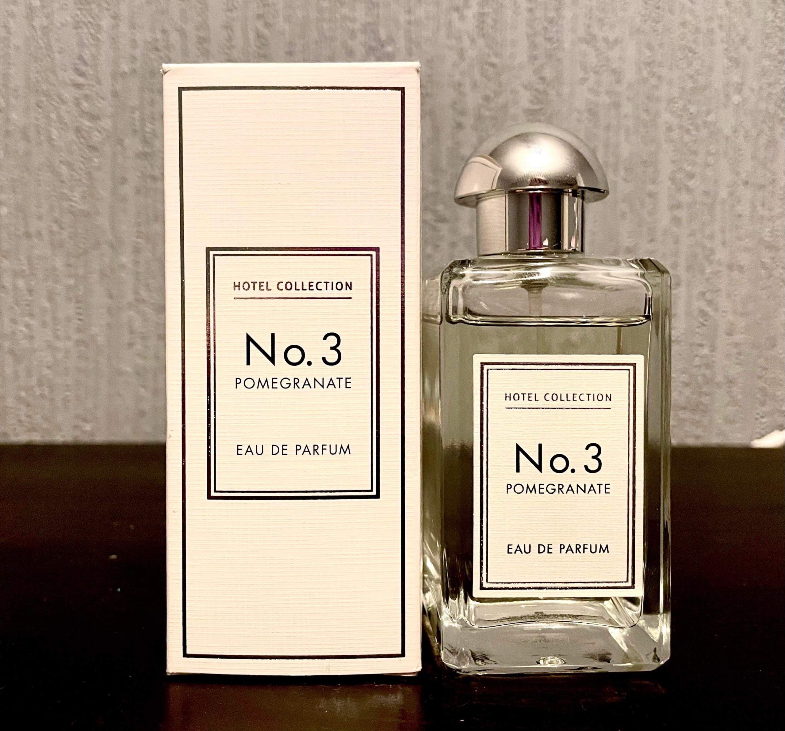 More Different And Intriguing Scents Of A Hotel | Aldi Hotel Collection Pomegranate Noir Eau De Parfum A tall rectangular clear glass bottle full of some clear liquid with a metallic silver lid and a white label that has Aldi Hotel Collection Pomegranate Noir Eau De Parfum written in bold black writing on it next to a tall rectangular white cardboard box that has Aldi Hotel Collection No.3 Pomegranate Noir Eau De Parfum written in bold black writing on it, on a white background.