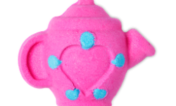 A small bright pink teapot shaped bath bomb that has a heart engraved on it and some bright gold lustre all over it, on a white background.