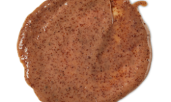 A medium circular puddle of some dark brown body scrub that has some pieces of Ground Almond Shells and Ground Date Stones running through it, on a white background.