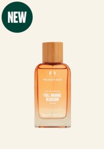 A tall rectangular clear glass bottle full of some translucent purple liquid with a light wooden Lid that has The Body Shop Full Orange Blossom Eau De Parfum written in bold white writing on it, on a white background.