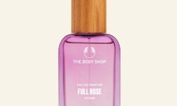 A tall rectangular clear glass bottle full of some translucent peach liquid with a light wooden Lid that has The Body Shop Full Rose Eau De Parfum written in bold white writing on it, on a white background.