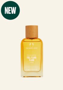 A tall rectangular clear glass bottle full of some translucent pink liquid with a light wooden Lid that has The Body Shop Full Ylang Ylang Eau De Parfum written in bold white writing on it, on a white background.