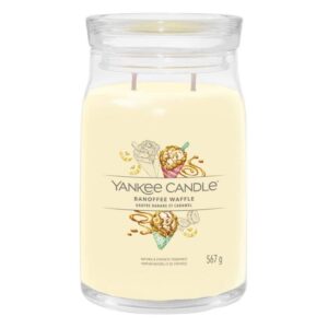 A tall cylindrical glass jar full of some creamy white wax with a label that has Yankee Candle written in white writing, Banoffee Waffle written in black writing, and a picture of some scoops of Banana Ice Cream and some pieces of some broken up waffle cone on it, on a white background..