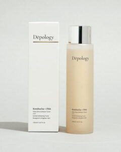 A tall cylindrical clear plastic bottle full of some creamy white liquid with a silver plastic lid that has Depology Daily Glow Exfoliant Boosting Toner written in bold black writing on it next to a tall rectangular white cardboard box that has Depology Daily Glow Exfoliant Boosting Toner written in bold black writing on it, on a white background.