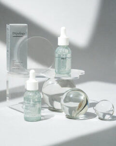 A short cylindrical translucent green glass bottle with a white rubber pipette lid that has Depology Dark Spot Correcting N.A.T Serum written in bright white writing on it next to a short cylindrical translucent green glass bottle with a white rubber pipette lid that has Depology Dark Spot Correcting N.A.T Serum written in bright white writing on it surrounded by some various different coloured glass balls, on a white background.
