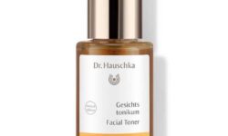 A short cylindrical clear glass bottle full of some clear liquid with a gold metal spray lid and a white label that has Dr. Hauschka Facial Toner written in bold black writing on it, on a white background.