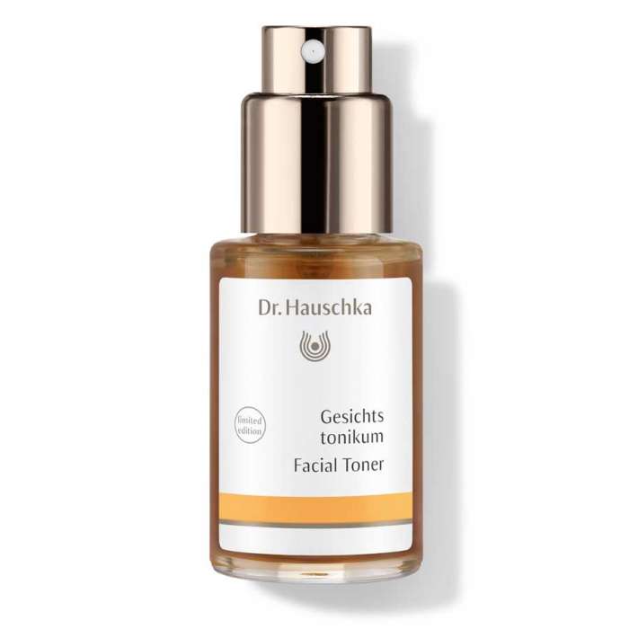 A short cylindrical clear glass bottle full of some clear liquid with a gold metal spray lid and a white label that has Dr. Hauschka Facial Toner written in bold black writing on it, on a white background.