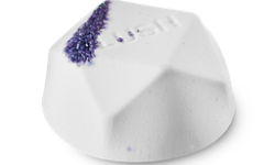 A medium bright white flat hexagonal shaped bath bomb that has some pieces of Demerara Sugar embedded in it, on a white background.