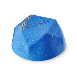 A medium dark blue flat hexagonal shaped bath bomb that has some pieces of Demerara Sugar embedded in it, on a white background.