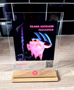 A tall rectangular piece of clear Acrylic that has a picture of a white man, a large bright red play button, and Black Sabbath Paranoid written in bright white writing on it stood on a rectangular light wooden stand, on a white background.