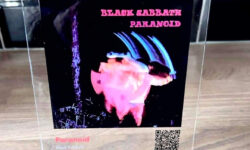 A tall rectangular piece of clear Acrylic that has a picture of a white man, a large bright red play button, and Black Sabbath Paranoid written in bright white writing on it stood on a rectangular light wooden stand, on a white background.