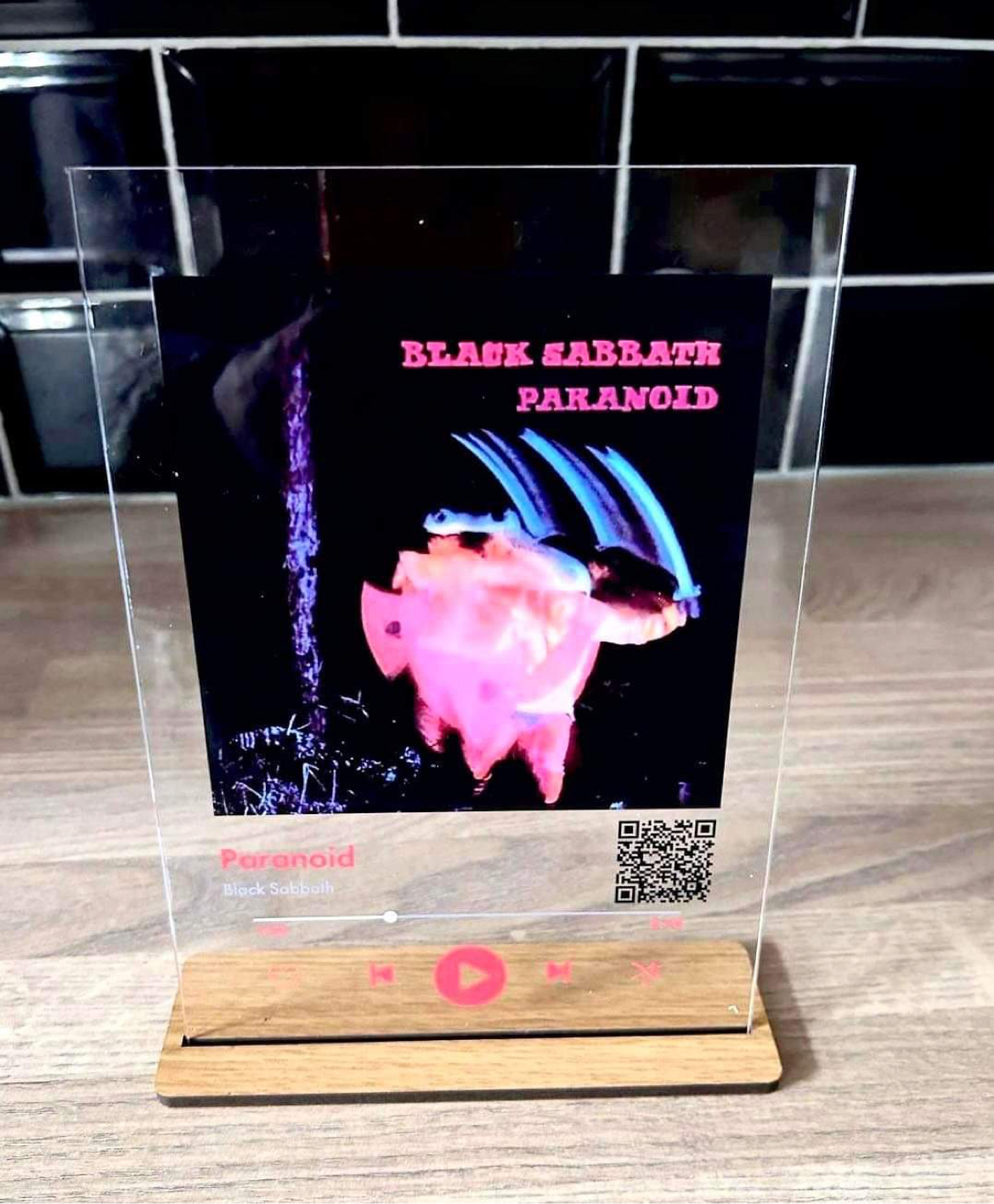 Me And My Love Of Music | Me And My Glass Personalised Spotify Glass Plaque | Gifted A tall rectangular piece of clear Acrylic that has a picture of a white man, a large bright red play button, and Black Sabbath Paranoid written in bright white writing on it stood on a rectangular light wooden stand, on a white background.