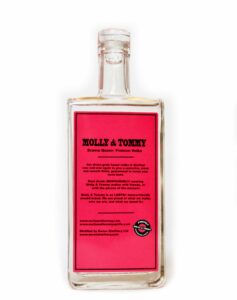 A large rectangular clear glass bottle full of some clear liquid with a clear plastic lid and a pink label that has Molly & Tommy Spirits Drama Queen Vodka written in bold black writing on it, on a white background.