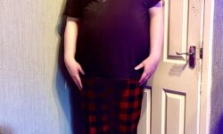 A tall bearded man wearing a bright red crew neck t-shirt, a pair of bright red pyjama pants with a black waistband that has Box Menswear written in bright white writing on it and a pair of grey and white checked socks stood against a white wooden wall.