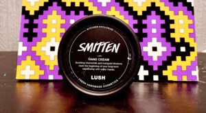 A short cylindrical black plastic tub with a black plastic lid and a black label that has Lush Smitten Hand Cream written in bright white writing on it, on a white background.
