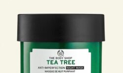 A short cylindrical clear glass jar full of some dark green liquid with a dark green plastic lid and a dark green label that has The Body Shop Tea Tree Anti-Imperfection Night Mask written in bright white writing on it, on a white background.