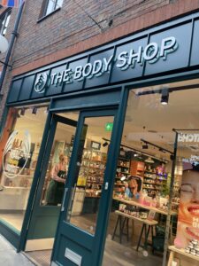 A large brown stone rectangular shaped building with a large rectangular dark green board over it's doors that has The Body Shop written in bold white writing on it, on a white background.