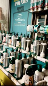 A collection of several large rectangular clear glass dispensers full of some various different coloured liquid next to a collection of some tall cylindrical silver aluminium bottles with some white Plastic pump lids and a large rectangular green board that has Refill Station written in bright white writing on it against a light grey wall.