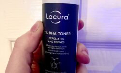 A short cylindrical clear plastic bottle with a black plastic lid and a light blue label that has Aldi Lacura 2% BHA Toner written in bright white writing on it, on a white background.